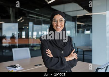 Porträt einer muslimischen Frau in Hijab bei der Arbeit im Büro, einer Geschäftsfrau, die lächelt und mit gekreuzten Armen auf die Kamera schaut. Stockfoto