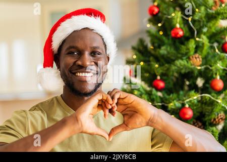 afroamerikanischer Mann, der eine Herzgeste mit Händen im Weihnachtshut in der Nähe des weihnachtsbaums zu Hause zeigt Stockfoto