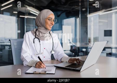 Arabische muslimische Frau in Hijab Arzt, der in einem modernen Klinikbüro mit Laptop arbeitet, Arzt auf Papier, in weißem medizinischen Mantel mit Stethoskop und Brille. Stockfoto