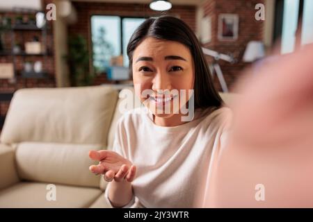 Glückliche junge asiatische Chineese filipino Frau sitzt auf dem Sofa zu Hause halten Telefon am Bildschirm winkende Hand Video Anruf Entfernung Freund online in mobilen Chat-App mit Smartphone-Videochat-Anwendung Stockfoto