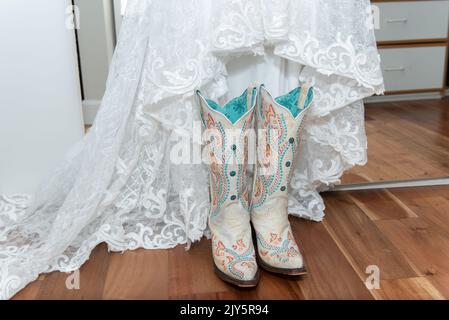 Weißes Spitzenkleid der Braut wird für eine westliche Hochzeitszeremonie mit weißen bestickten Cowgirl-Stiefeln kombiniert. Stockfoto