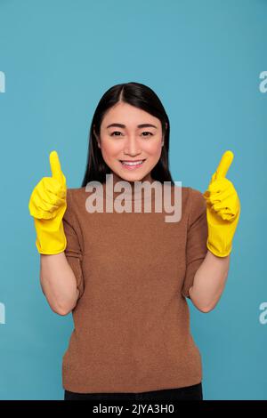 Aufgeregt glücklich und fröhlich Zimmermädchen von der Hausarbeit Daumen nach oben und tragen gelbe Handschuhe für Handschutz isoliert auf einem blauen Hintergrund, Reinigung Home Konzept, zufriedene Lichtung weiblich mit positiver Stimmung Stockfoto