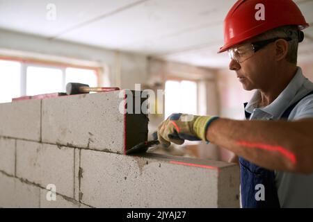 Maurer trägt Klebstoff auf autoklavierte, belüftelte Betonblöcke mit gekerbter Kelle auf. Der Auftragnehmer des Maurerarbeiters entfernt überschüssigen Mörtel aus Stockfoto