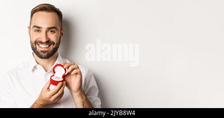 Nahaufnahme des glücklichen, gutaussehenden Mannes, der einen Vorschlag macht, den Ehering in der Box hält und lächelt, ihn bittet, ihn zu heiraten, weißer Hintergrund Stockfoto