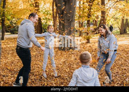 Eine junge Familie von vier Mitgliedern freut sich in einem Wald und wirft an einem Herbsttag Blätter in die Luft. Herbstlaub. Spaß für die ganze Familie im Herbst. Kleinkind Stockfoto
