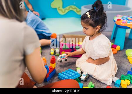 Spielerisch im Kindergarten lernen. Kleinkind kleines Mädchen und die Lehrerin spielen mit bunten Kunststoff-Spielhäusern, Bausteinen, Autos und Booten. Entwicklung von Phantasie, Kreativität, Feinmotorik und grobmotorischen Fähigkeiten. Hochwertige Fotos Stockfoto