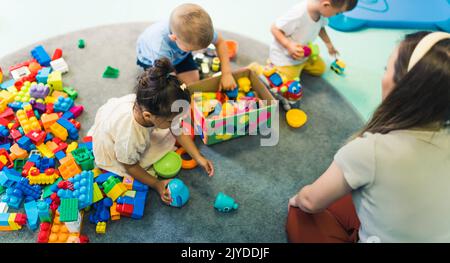 Spielzeit im Kindergarten. Kleinkinder mit ihrem Lehrer sitzen auf dem Boden und spielen mit Bausteinen, bunten Autos und anderen Spielzeugen. Hochwertige Fotos Stockfoto