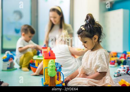 Spielerisch im Kindergarten lernen. Kleinkind kleines Mädchen und die Lehrerin spielen mit bunten Kunststoff-Spielhäusern, Bausteinen, Autos und Booten. Entwicklung von Phantasie, Kreativität, Feinmotorik und grobmotorischen Fähigkeiten. Hochwertige Fotos Stockfoto