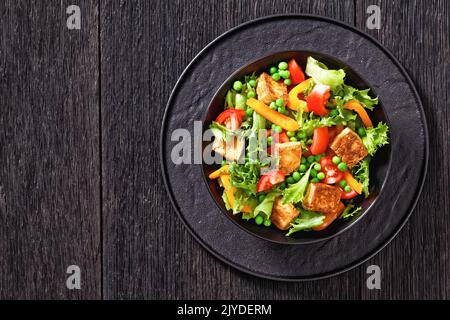 Tofu-Salat mit knackigen Salatblättern, grünen Erbsen, Tomaten, gelber Paprika in schwarzer Schüssel auf Holztisch, horizontale Ansicht von oben, flach liegend, Stockfoto