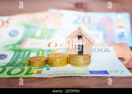 Miniaturhausmodell mit Münzstapeln und Euro-Banknoten auf einem Holztisch, selektiver Fokus. Home Loan-Konzept. Stockfoto