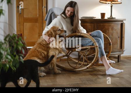 Junge Frau mit Hund Golden Retriever, Katze sitzt auf Schaukelstuhl. Gemütliche Atmosphäre, langsames Leben, Menschen mit Haustieren. Interieur im Vintage-Haus Stockfoto