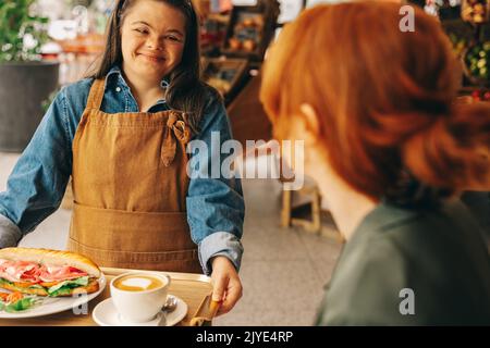 Fröhliche Kellnerin mit Down-Syndrom serviert einem Gast ein Sandwich und Kaffee in einem trendigen Café. Freundliche Frau mit einer intellektuellen Behinderung bei der Arbeit Stockfoto