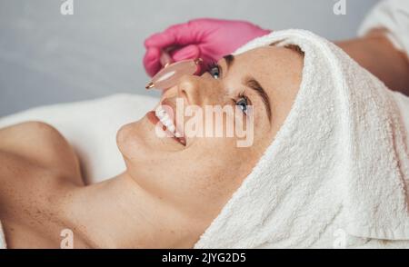 Seitenansicht einer Frau, die auf einem Massagebett liegt und in einem Schönheitssalon eine Gesichtsmassage erhält. Moderne Beauty-Spa-Behandlung und Relax-Concep. Stockfoto