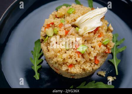 Couscous Porridge mit Gemüse, Butter und Kräutern, auf einem Teller, auf dunklem Grund Stockfoto