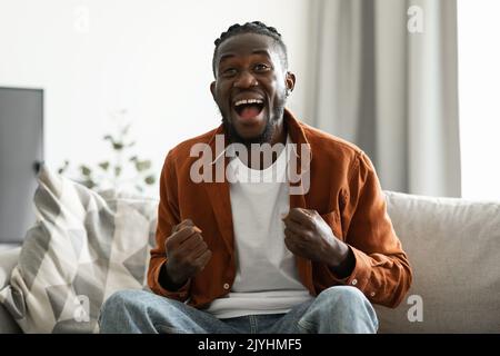 Emotionaler afroamerikanischer Mann schreit und gestikiert JA in Freude, zu Hause auf dem Sofa sitzend, Raum kopieren Stockfoto