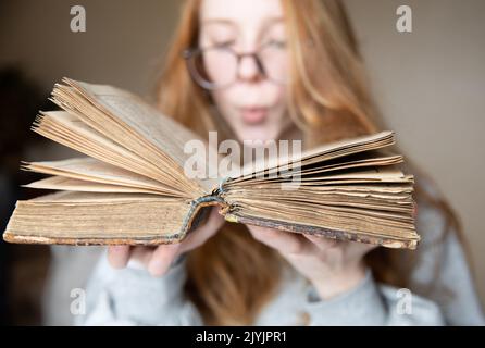 Ein offenes Vintage-Buch in den Händen eines niedlichen Teenagers mit Brille und roten Haaren, in einem halbdunklen Raum, der vom Licht des Fensters beleuchtet wird Stockfoto
