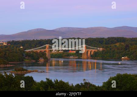 Die Menai Suspension Bridge in der Dämmerung mit den Snowdonia Mountains im Hintergrund. Anglesey, Nordwales, Großbritannien. Stockfoto