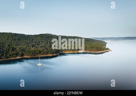 Luftdrohnenaufnahme der Adriatischen Insel am frühen Morgen. Nadelwald und türkisfarbenes, kristallklares Meerwasser.Ein Segelboot, das auf ruhigem Wasser vor Anker liegt. Stockfoto