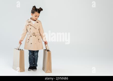 In voller Länge stilvolles Kind in Trenchcoat und Jeans mit Einkaufstaschen auf grauem, schwarzem freitagskonzept, Stockbild Stockfoto
