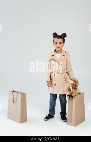 In voller Länge von stilvollen Kind in Trenchcoat und Jeans halten Einkaufstasche mit Spielzeug Pferd auf grau, Stockbild Stockfoto