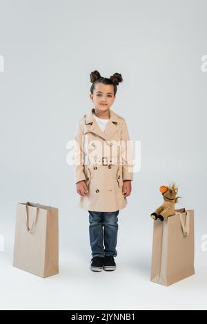 In voller Länge fröhliche Kind in Trenchcoat und Jeans stehen in der Nähe von Einkaufstaschen mit weichem Spielzeug auf grau, Stock Bild Stockfoto