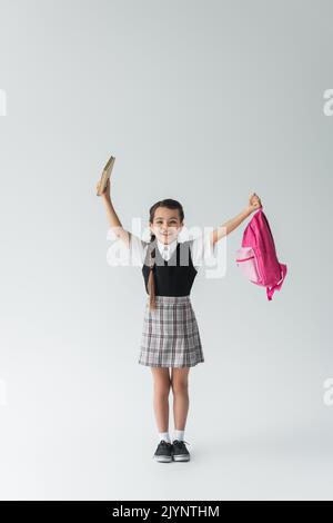 Volle Länge der aufgeregt Schulmädchen in Uniform halten rosa Rucksack und Buch auf grau, Stock Bild Stockfoto