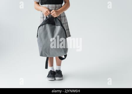 Beschnittene Ansicht von Schulmädchen in Uniform halten Rucksack und stehen auf grau, Stock Bild Stockfoto