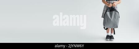 Beschnittene Ansicht von Schulmädchen in Uniform halten Rucksack und stehen auf grau, Banner, Stock Bild Stockfoto