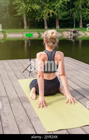 Eine Frau sitzt im Sommer auf einer hölzernen Plattform, macht Yoga auf einer grünen Matte am Teich Stockfoto