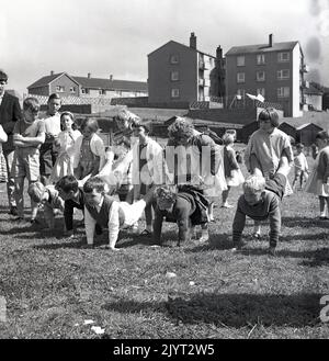 1965, historischer, n. queensferry Gala-Tag, Kinder bereit, eine lustige Aktivität zu starten, ein Rad-Schubkarren-Rennen draußen auf dem Gras auf einem Feld eines Wohnguts in North Queensferry, Fife, Schottland, Großbritannien, die tapferen jungen Mädchen mit den Beinen der Jungen! Stockfoto