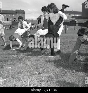 1965, historischer Gala-Tag im Norden von queensferry, Jugendliche, die draußen auf dem Rasen auf einem Feld eines Wohnguts in North Queensferry, Fife, Schottland, Großbritannien, an einem Rennen mit dem Schubkarren teilnehmen, die Mädchen halten die Beine der Jungen! Stockfoto