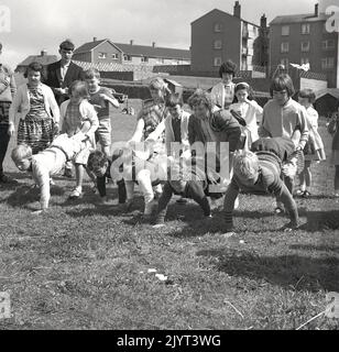 1965, historischer, n. queensferry Gala-Tag, Kinder bereit, ein Spiel zu starten, ein Rennen mit dem Schubkarren draußen auf dem Gras auf dem Gelände eines Wohnguts in North Queensferry, Fife, Schottland, Großbritannien, die jungen Mädchen halten die Beine der Jungen! Stockfoto