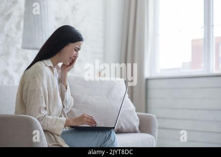 Eine junge verärgerte Frau kommuniziert online per Videoanruf von einem Laptop aus mit einem Psychologen. Sie sitzt zu Hause auf dem Sofa am Fenster, weint, wischt ihre Tränen mit einer Serviette ab. Ich fühle mich deprimiert, traurig. Stockfoto