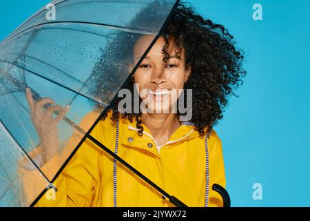 Porträt einer afrikanischen Frau mit weißem Lächeln in gelbem Regenmantel mit transparentem Regenschirm und Wassertropfen auf blauem Hintergrund. Herbstsaison kontra Stockfoto