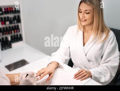 Schöne Frau im Bademantel während der Maniküre im Spa-Salon, kümmert sich um ihre Nägel. Manikuristin bedeckt Frauennägel mit Nagellack Stockfoto