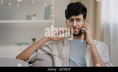 Bärtiger Mann mit Brille sitzt auf dem Sofa zu Hause im Gespräch auf Handy-Gespräch mit Freunden Freundin freundlich Remote-Gespräch Stockfoto