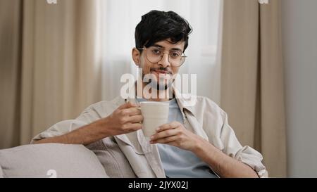 Arabisch bärtig Millennial Mann latein Mann in Gläsern sitzen zu Hause genießen köstliche heiße Früchte Kräutertee Kaffee trinken Kakao trinken Cappuccino Stockfoto