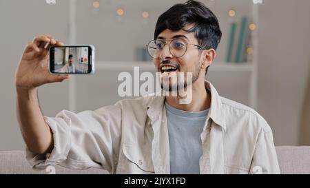 Junge fröhlich arabischen Kerl bärtigen Mann trägt Brille sitzen zu Hause drinnen Couch mit Handy Kamera Aufnahme Video Blog männlichen Blogger Stockfoto