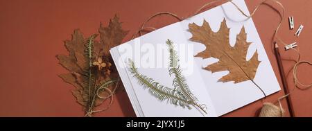 Getrocknetes gepresstes Blatt auf Papier aus Notizbuch mit Bleistift, Schere und Garn auf braunem Hintergrund. Hobby, Handarbeit, Blumenkunst und Boho-Konzept. Banner Stockfoto