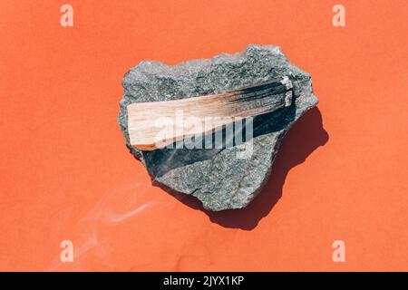 Schwelender palo santo-Stock mit Rauch auf Steinständer auf orangerotem Hintergrund, brennender Weihrauch. Flache Lay-Komposition mit heiligen Holzstäben für medi Stockfoto