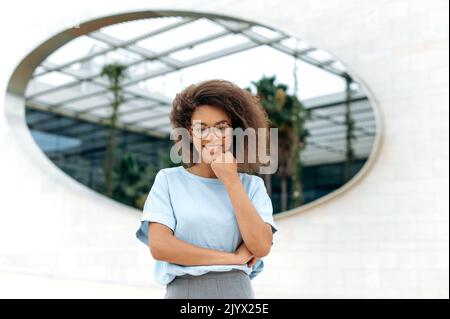 Porträt einer schönen selbstbewussten, eleganten afroamerikanischen Frau mit lockigen Haaren, in einem blauen Hemd, mit Brille, im Freien stehend, auf die Kamera schauend, freundlich lächelnd Stockfoto