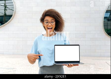 Aufgeregt erstaunt afroamerikanisch lockig haarige junge Frau, steht im Freien, hält einen offenen Laptop mit weißem Mock-up-Bildschirm in der Hand, zeigt den Finger auf ihn, sieht überrascht auf die Kamera Stockfoto