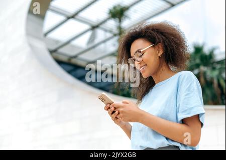 Online-Chat. Positive afroamerikanische junge Frau mit lockigen Haaren, stilvoll gekleidet, mit ihrem Smartphone im Freien stehend, Messaging mit Freunden in sozialen Netzwerken, beantwortet E-Mails und lächelt Stockfoto