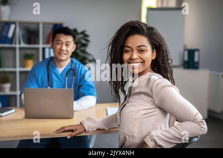 Lächelnd Erwachsene asiatische männliche Arzt mit Laptop Beratung junge afroamerikanische Frau Patientin, Blick in die Kamera Stockfoto