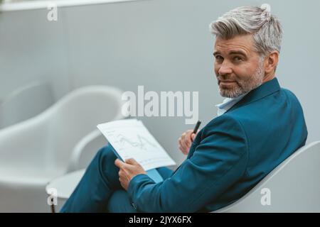 Vielbeschäftigter reifer Geschäftsmann arbeitet mit Dokumenten, während er im modernen Coworking sitzt Stockfoto