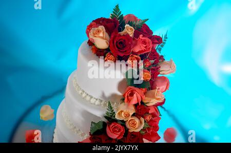 Hochzeitstorte mit echten Rosen dekoriert. Kaskadierung von roten und rosafarbenen Rosen auf einem eleganten dreistufigen weißen Milchkuchen. Hintergrund ist ein verträumtes tiefes Blau Stockfoto