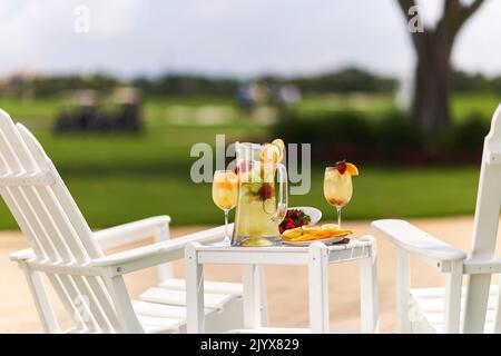 Limonade im Glaskrug mit Orangen in Scheiben und Limetten. Garniert mit Erdbeeren. Draußen auf einem kleinen Tisch mit zwei weißen Adirondack-Stühlen. Stockfoto