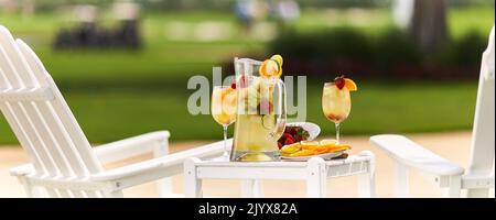 Limonade im Glaskrug mit Orangen in Scheiben und Limetten. Garniert mit Erdbeeren. Draußen auf einem kleinen Tisch mit zwei weißen Adirondack-Stühlen. Stockfoto