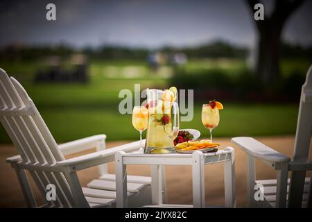 Limonade im Glaskrug mit Orangen in Scheiben und Limetten. Garniert mit Erdbeeren. Draußen auf einem kleinen Tisch mit zwei weißen Adirondack-Stühlen. Stockfoto