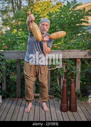 Der ältere Mann (Ende 60s) trainiert mit indischen Clubs und schweren persischen Meels auf seiner Terrasse im Hinterhof, funktionelles Fitnesskonzept mit traditionellem ebenfalls Stockfoto